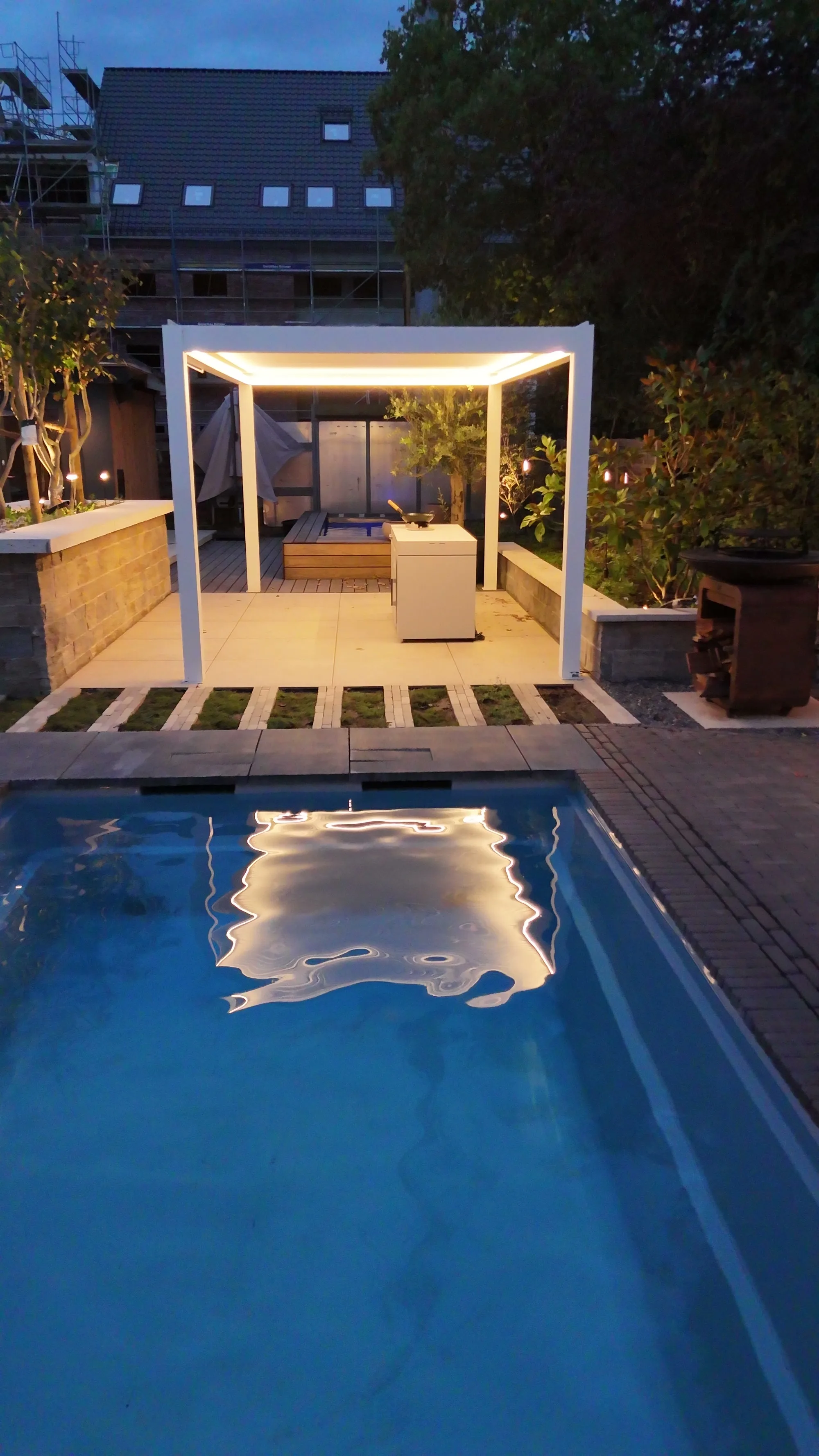 Weiße Pergola mit Lamellendach, am Rand läuft ein warmes Lichtband, davor junge Bäume.